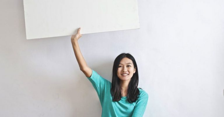 Premium Real Estate - Cheerful Asian woman sitting cross legged on floor against white wall in empty apartment and showing white blank banner